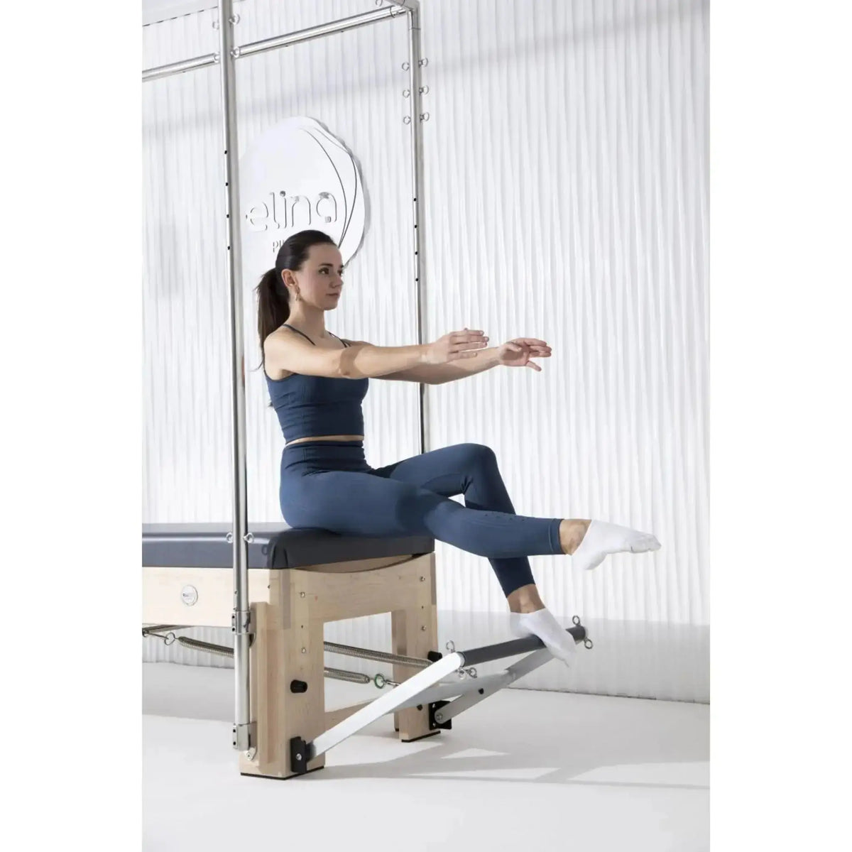 Elina Pilates Cadillac Elite Reformer aus Rock-Ahorn – Frau sitzt am Fußende mit angehobenen Beinen im Studio