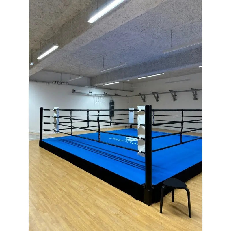 SportCom Pro-Trainingsboxring mit 30 cm Podium, mit blauem Boden auf Holzboden, Hocker in der Ecke