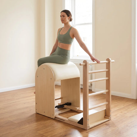 Frau sitzt seitlich auf dem Fass des Elina Pilates Elite Ladder Barrel, eine Hand auf der Sprosse