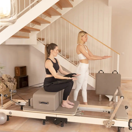 Elina Pilates Klappbarer Reformer aus Ahornholz, zwei Frauen trainieren mit Sitzbox auf dem Reformer im Wohnbereich