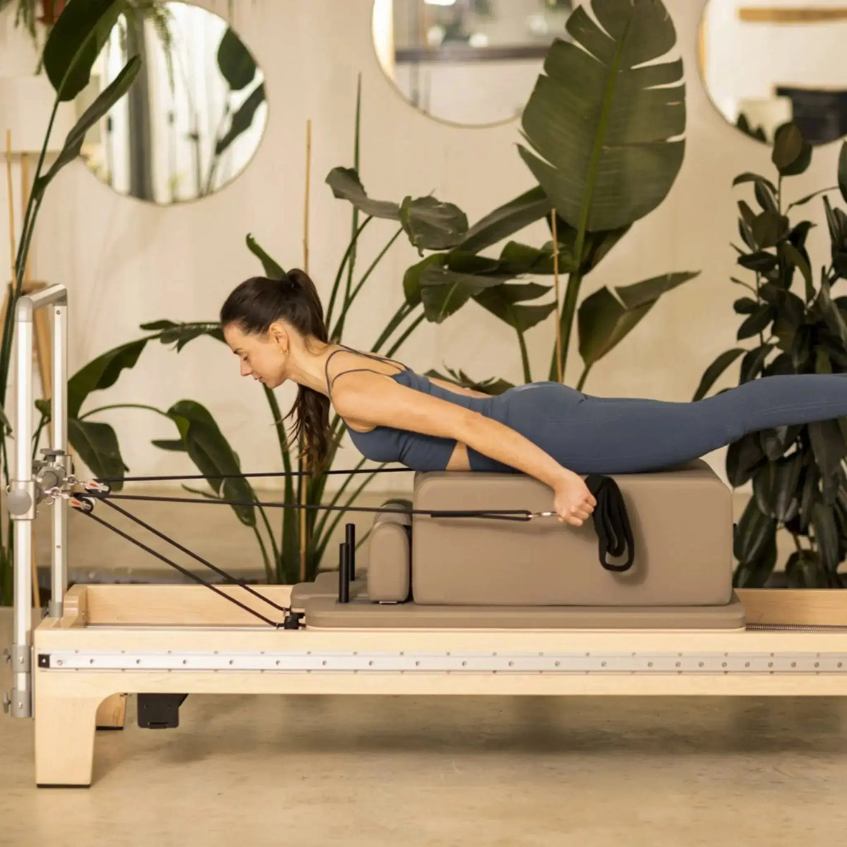 Elina Pilates Master Instructor Reformer aus Ahornholz – Frau trainiert in Bauchlage auf dem Schlitten im hellen Studio mit Pflanzen