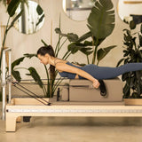 Elina Pilates Master Instructor Reformer aus Ahornholz – Frau trainiert in Bauchlage auf dem Schlitten im hellen Studio mit Pflanzen