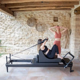 Elina Pilates Nubium Black Edition Reformer aus schwarzem Aluminium im Steinraum, Frau trainiert liegend mit Schlaufen