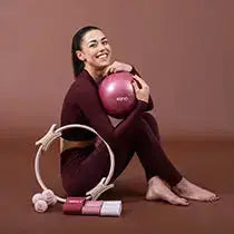 Elina Pilates Pilates-Ring in Rosa, lächelnde Frau sitzt mit Ring und rosa Pilates-Ball vor braunem Hintergrund