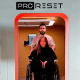 Frau im Rollstuhl im Innenraum der ProReset Matrix Hyperbarkammer, Mann steht hinter ihr, rote LED