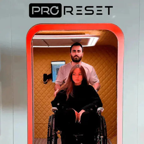 Frau im Rollstuhl im Innenraum der ProReset Matrix Hyperbarkammer, Mann steht hinter ihr, rote LED