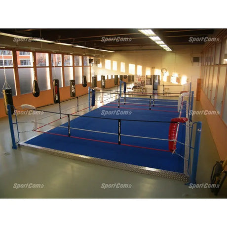 SportCom Ecofix Doppel-Boxring mit Aluminiumrand, in einer Halle mit Holzdecke und Boxsäcken, blauer Boden