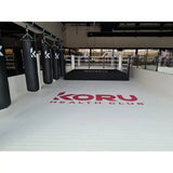 SportCom Flexi-Roll Ringermatte von Dollamur, weiße Fläche mit rotem Logo vor Sandsäcken und Boxring
