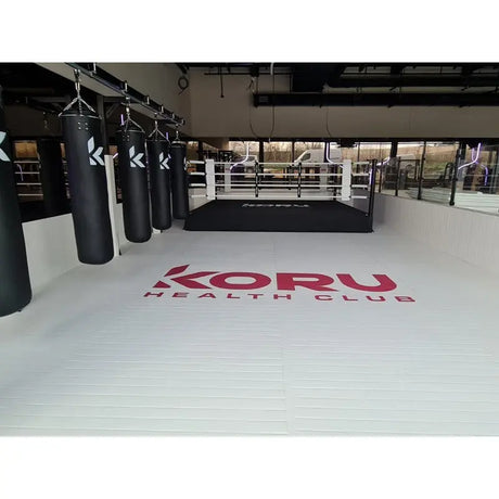 SportCom Flexi-Roll Ringermatte von Dollamur, weiße Fläche mit rotem Logo vor Sandsäcken und Boxring