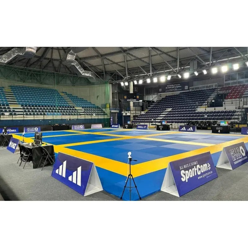 SportCom Judo-Tatami Reisstroh 2 × 1 m, 5 cm, glatte Unterseite, blau-gelbe Wettkampffläche in einer Arena mit Tribünen