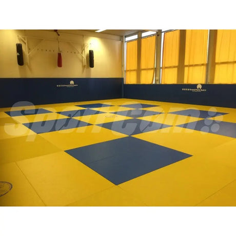SportCom Judo-Tatami Reisstroh 2 × 1 m, 5 cm, glatte Unterseite, gelbe Fläche mit blauem Schachbrettmuster in einer Halle