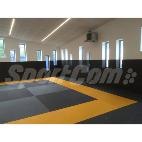 SportCom Judo-Tatami Reisstroh 2 × 1 m, 5 cm, glatte Unterseite, graues Schachbrettmuster mit gelbem Rand in Halle mit hohen Fenstern