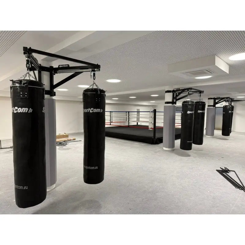 SportCom Sandsack-Wandhalterung mit Schiene und Laufwagen mit schwarzen Sandsäcken in einer Halle mit Boxring