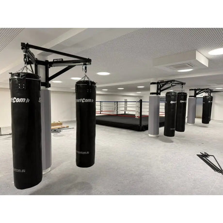 SportCom Sandsack-Wandhalterung mit Schiene und Laufwagen mit schwarzen Sandsäcken in einer Halle mit Boxring
