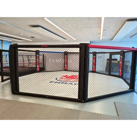 SportCom Wettkampf-MMA-Käfig 7 m auf Podium, Frontansicht mit roten Polstern und FMMAF-Matte in der Halle