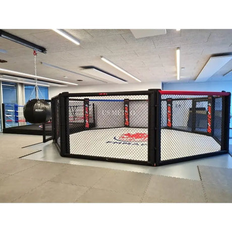 SportCom Wettkampf-MMA-Käfig 7 m auf Podium, in einer Trainingshalle mit Boxsäcken im Hintergrund